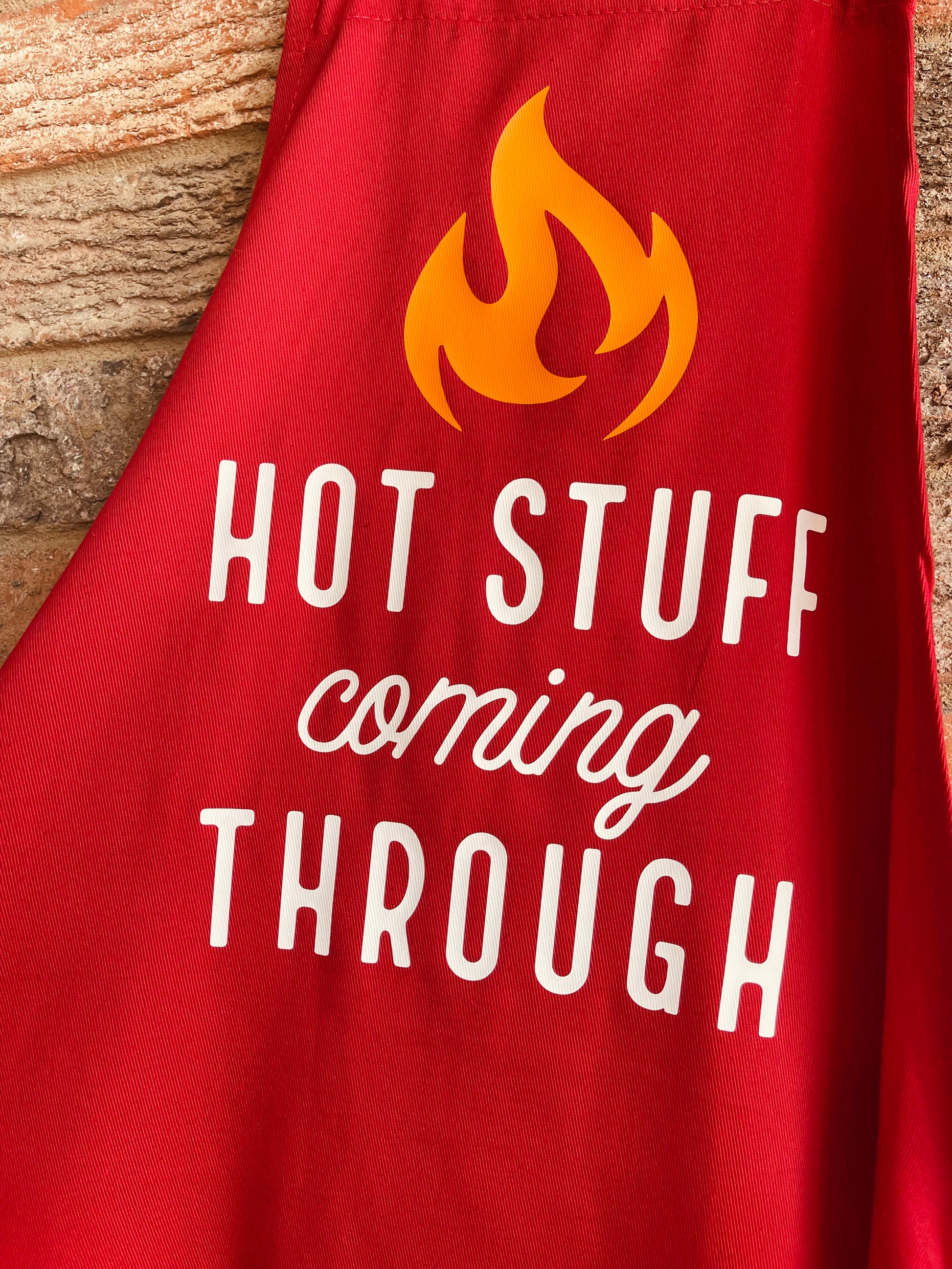 This is a red apron with a functional, adjustable tie that goes around the waist. It has "hot stuff coming through" in white lettering with a yellow flame above the design. 